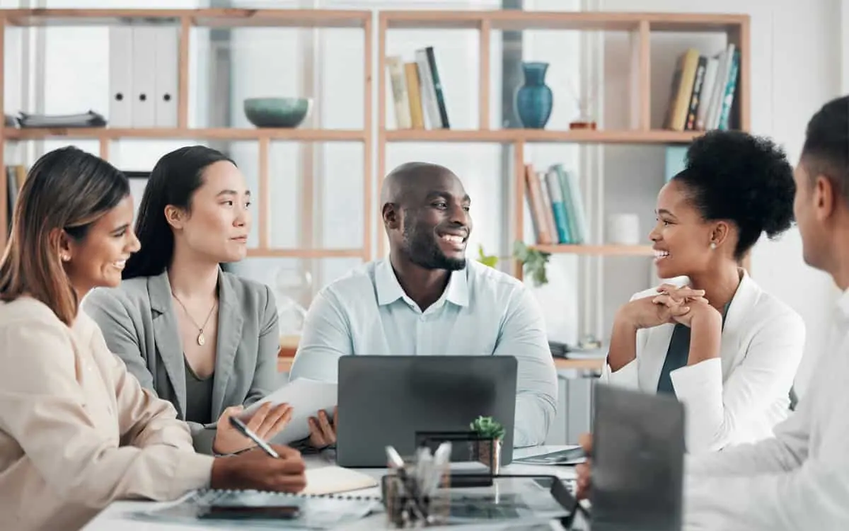 A diverse group of five people engaged in a meeting in a modern office, discussing services with notebooks, a laptop, and various office supplies on the table.