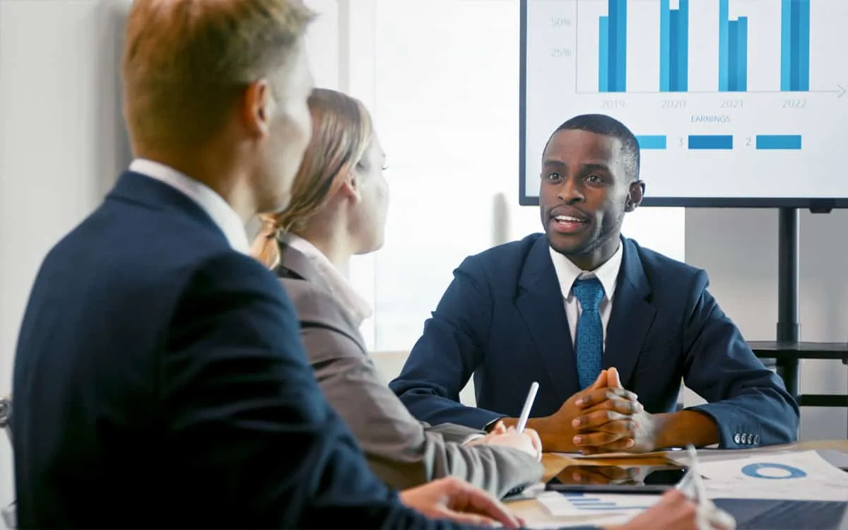 Three people in business attire are having a discussion in a meeting room, with a presentation screen displaying bar charts in the background, focusing on strategic financial solutions.