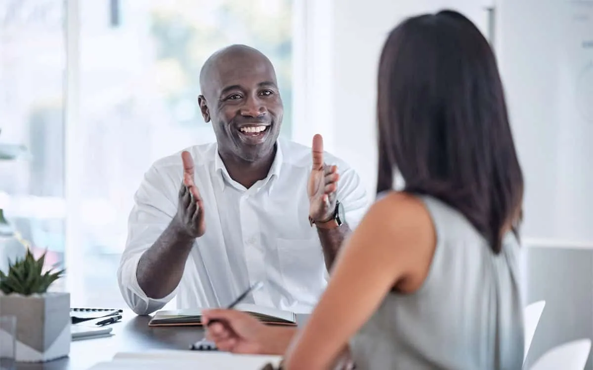 A man in a white shirt is smiling and gesturing with his hands while discussing bookkeeping with a woman with long hair sitting across from him in an office setting.