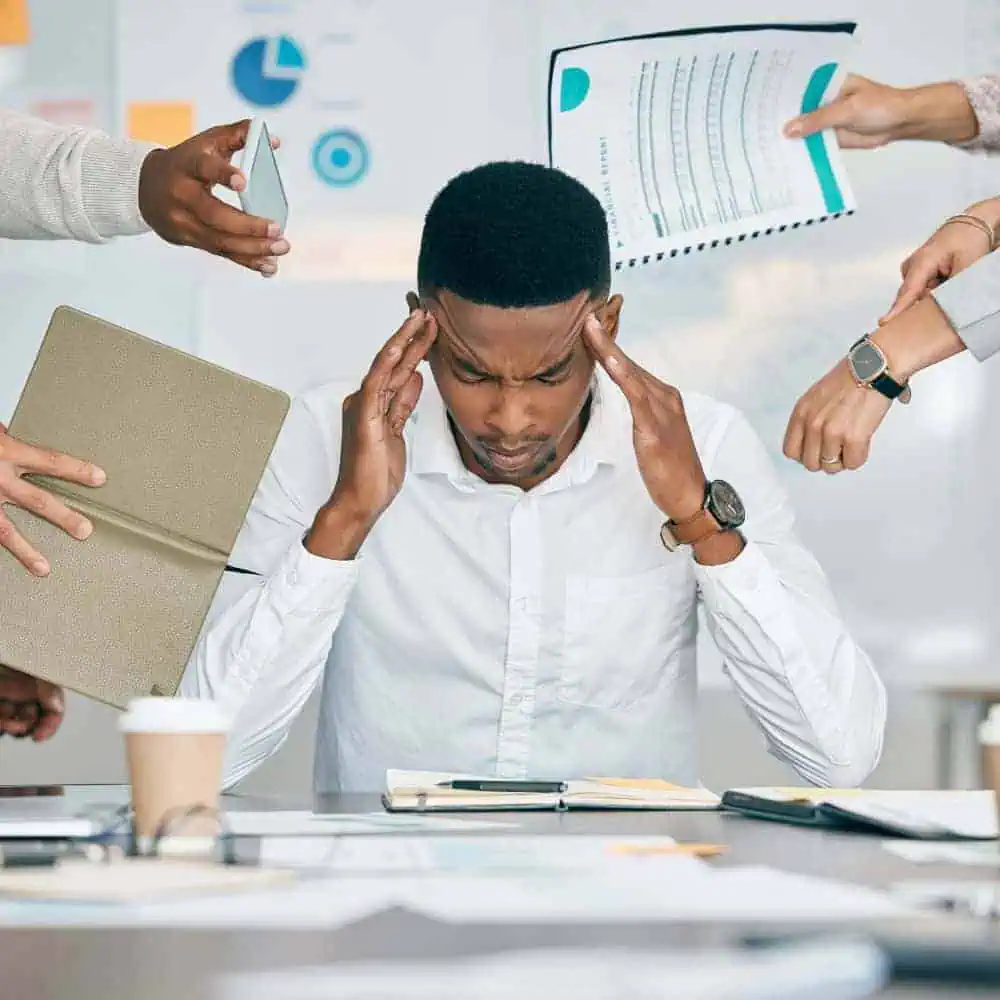 A man in a white shirt sits at a desk with a stressed expression, holding his head as multiple hands present documents, a phone, and a tablet to him—clearly in need of strategic financial solutions.