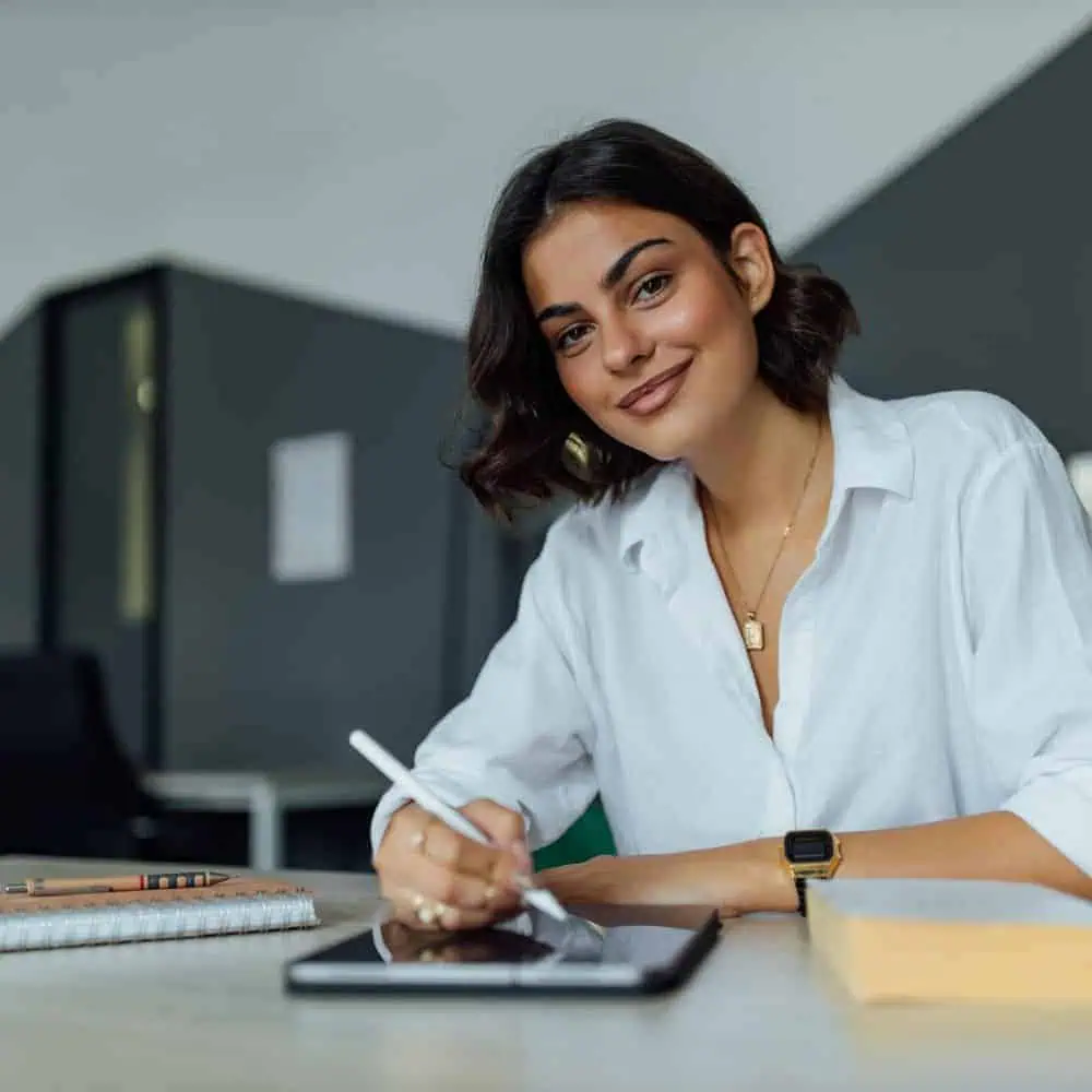 A person in a white shirt uses a stylus on a tablet at a desk, surrounded by notebooks and pencils, focusing intently on strategic financial solutions.