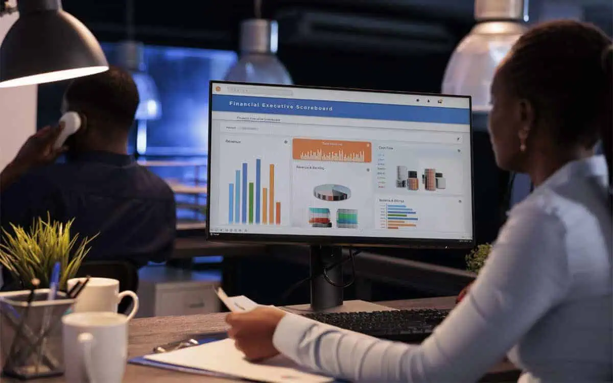 A person sits at a desk, engrossed in financial charts and graphs on the computer screen, indicating project finance analysis, while another individual talks on a phone in the background.