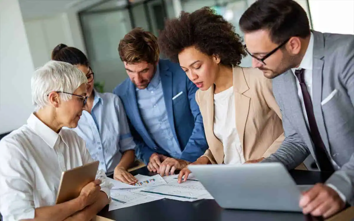 Five individuals in business attire collaborate around a table, examining documents and charts related to AR & AP Management. One person holds a tablet, and another uses a laptop.