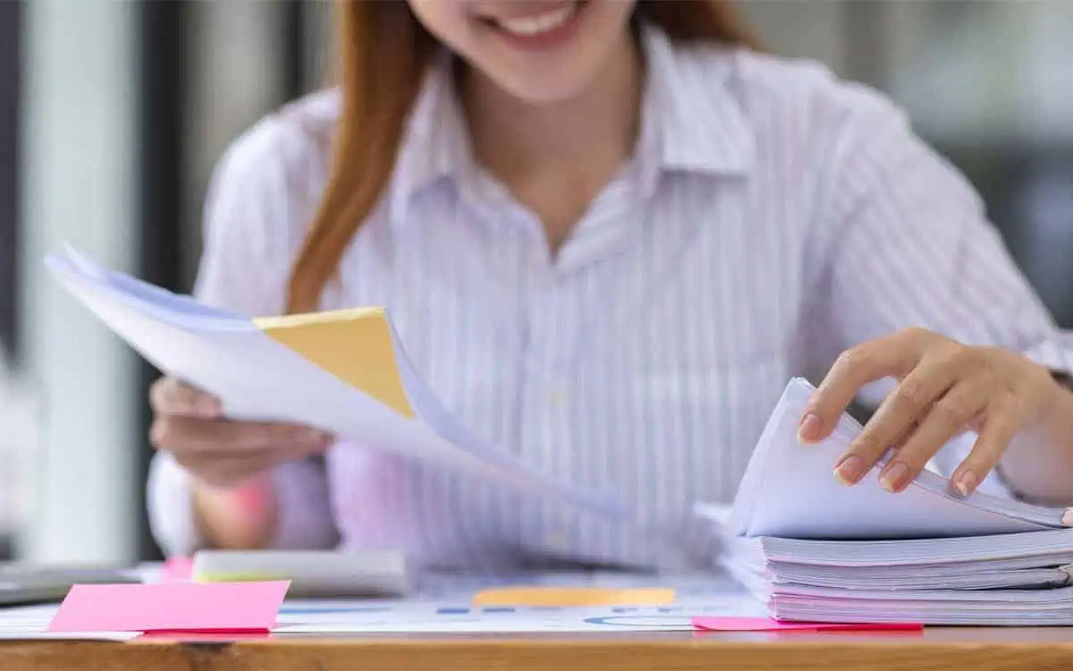 A person in a striped shirt is organizing a stack of papers on a desk with colorful sticky notes, meticulously sorting through Payroll documents and HR files.