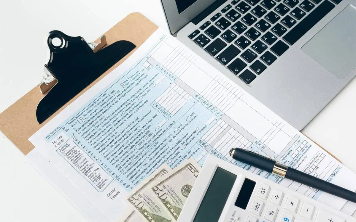 A tax form on a clipboard, a calculator, pen, dollar bills, and a laptop on a white desk—everything you need to explore our professional tax services.