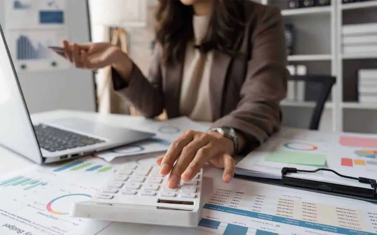 A person uses a calculator at a desk cluttered with financial charts and graphs, meticulously handling bookkeeping tasks while holding a credit card and working on a laptop.