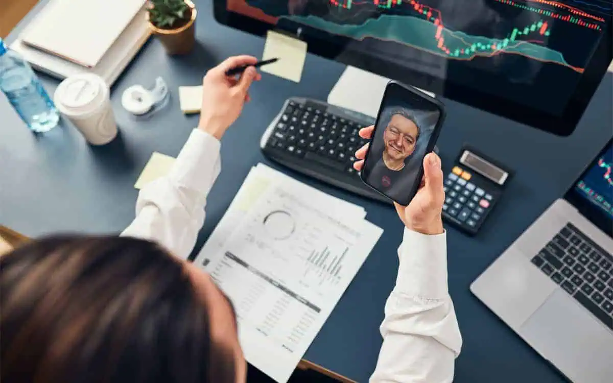 A person at a desk with financial documents and a computer displaying stock charts, holding a smartphone showcasing a video call with their virtual CFO.