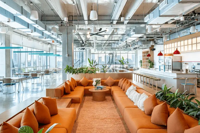 A modern, open-concept office space ideal for small businesses, featuring a large orange sectional sofa, a communal kitchen area, high ceilings, and large windows letting in plenty of natural light.