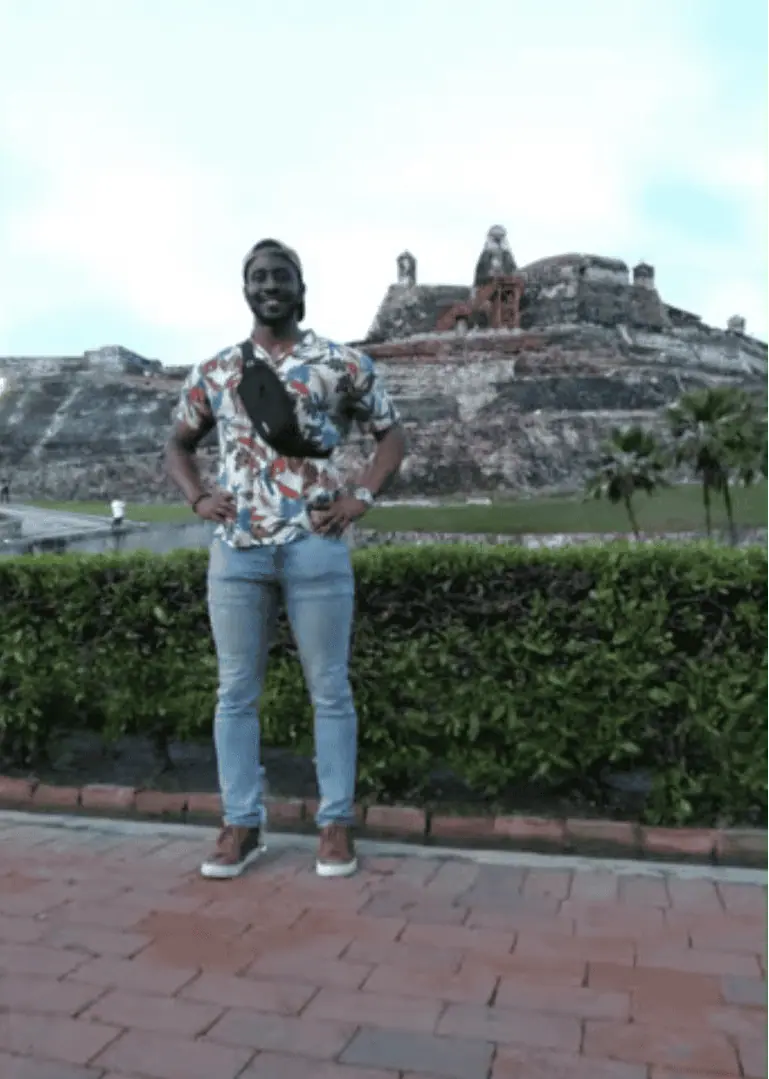 A person in a floral shirt and jeans stands with hands on hips in front of a historic fort structure surrounded by grass and greenery, showcasing the charm of an area that supports vibrant small businesses.