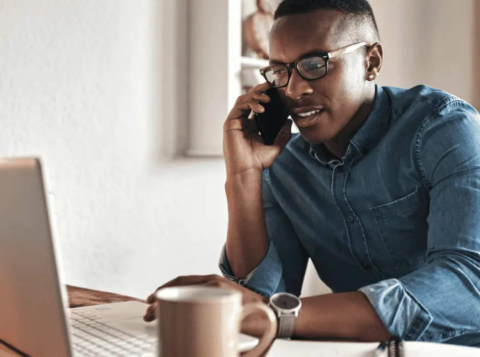 A person wearing glasses and a denim shirt is sitting at a desk, talking on the phone, and looking at a laptop. A coffee mug and a notebook are placed on the desk, suggesting they might be managing tasks for small businesses.