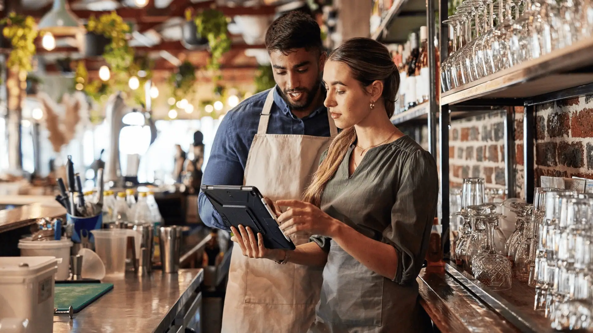 Two people stand behind a restaurant bar counter, looking at a tablet together. The man in an apron and the woman in a gray shirt seem focused as they review payroll & HR details. Glasses and other bar equipment are visible around them.