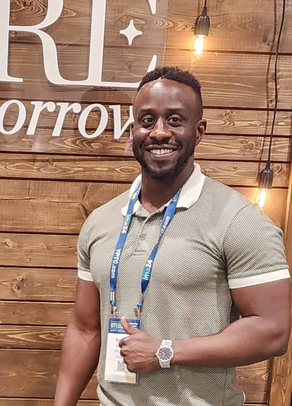 A man wearing a grey polo shirt with a conference badge and lanyard smiles and gives a thumbs up in front of a wooden wall adorned with hanging lights, signaling his support for small businesses.