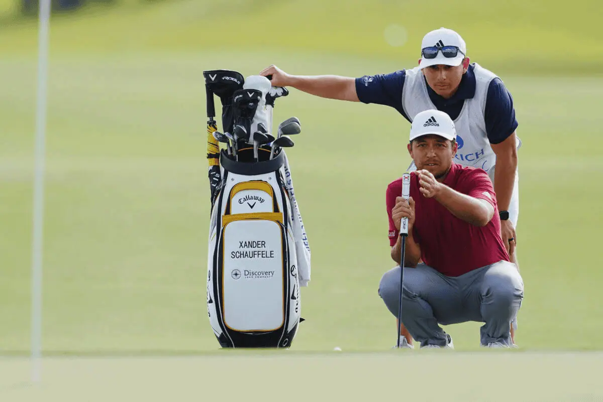 Golfer in a red shirt kneeling and aligning his putt on the green, with a caddie standing behind holding a golf bag labeled "Xander Schauffele," like a virtual CFO aligning financial strategies for success.