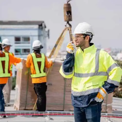 Construction worker in a high-visibility jacket and hard hat talks on a radio about project finance. Other workers, also in safety gear, move a large pallet in the background.