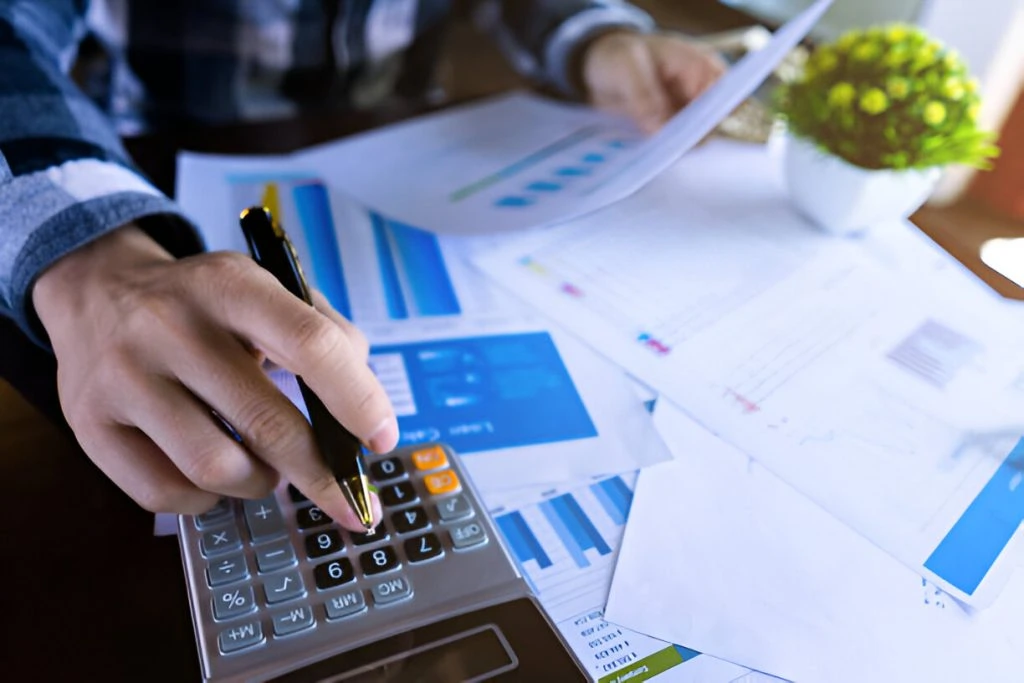 A person uses a calculator and holds a pen, with various financial documents and charts spread on a desk—illustrating what does a bookkeeper do for a small business by managing finances and keeping accurate records.