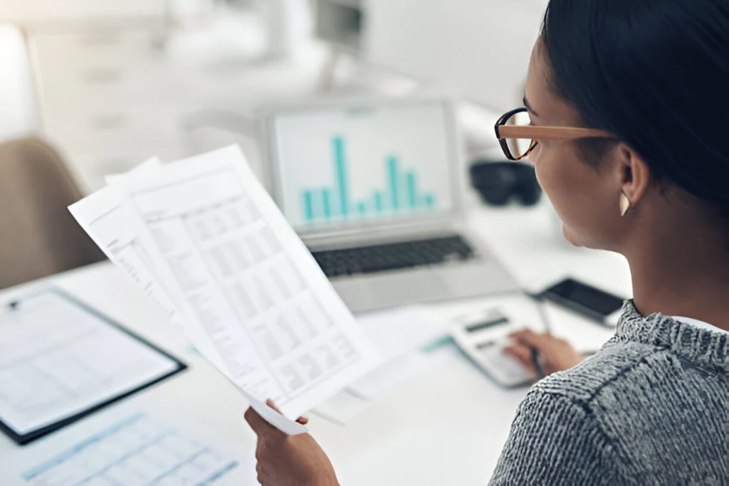 A person reviews printed financial documents and calculates bookkeeping cost for small business, with a laptop displaying bar graphs in the background.