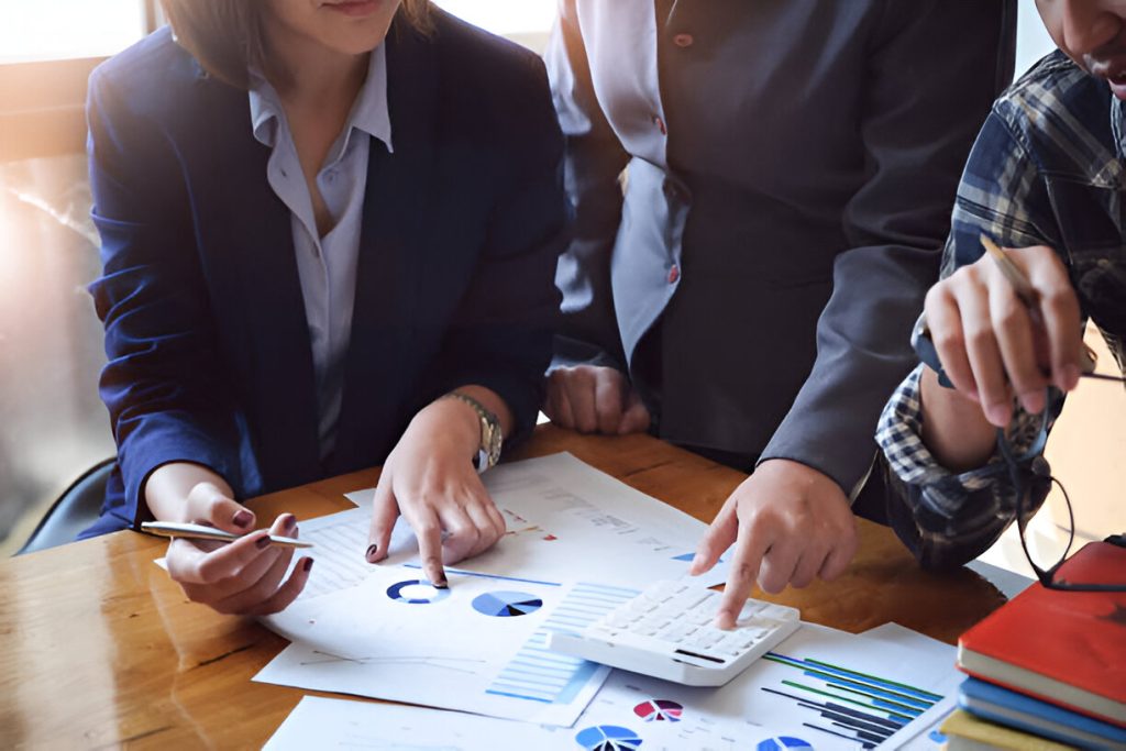 Three people in business attire review printed charts and use a calculator at a desk, discussing financial data while evaluating the best bookkeeping service for small business needs.