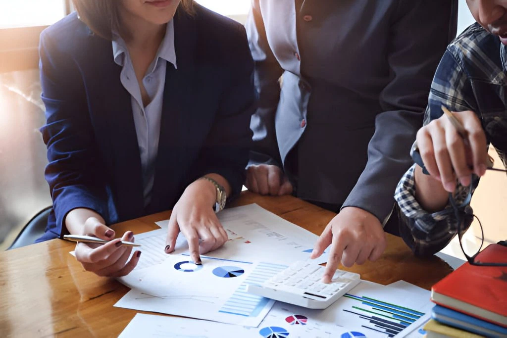 Three people in business attire review printed charts and use a calculator at a desk, discussing financial data while evaluating the best bookkeeping service for small business needs.