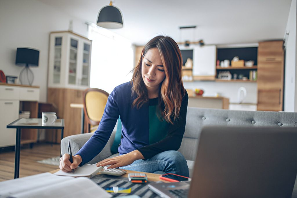 Woman sitting on a couch in a modern living room, writing in a notebook with her laptop, smartphone, and paperwork—perhaps calculating bookkeeping cost for small business—on the table in front of her.