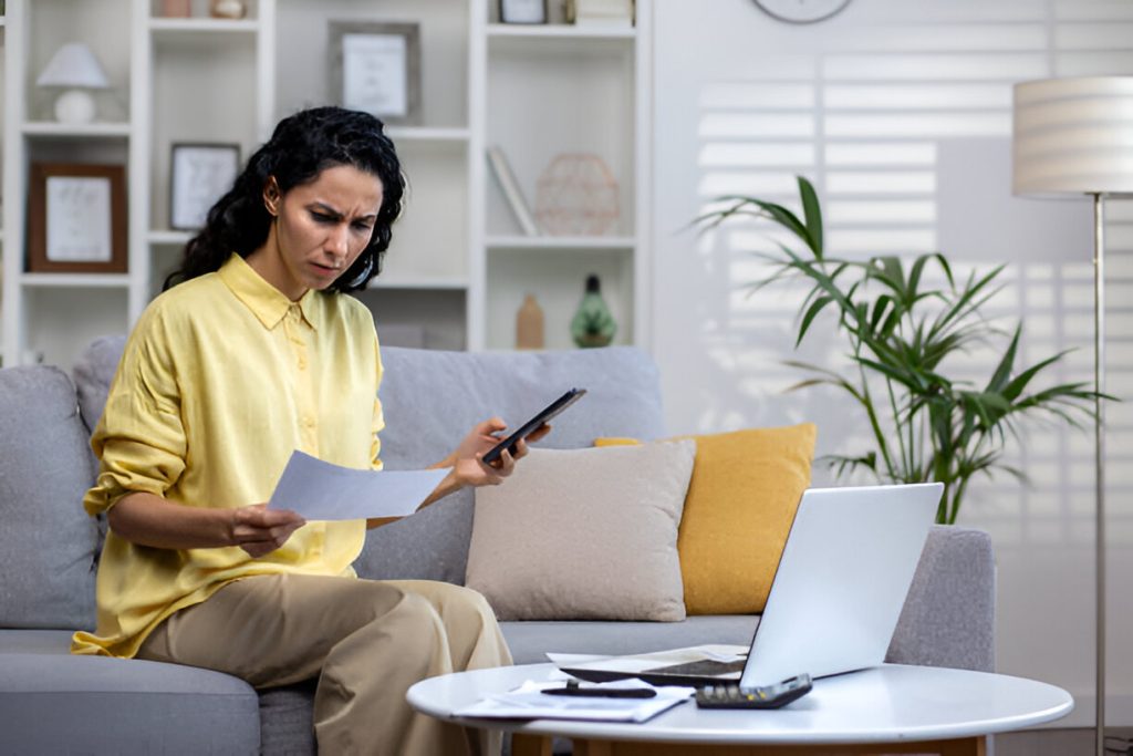 A woman sits on a couch holding papers and a phone, looking concerned—possibly worried about bookkeeping mistakes small business owners often face—with a laptop, calculator, and more papers on the coffee table in front of her.