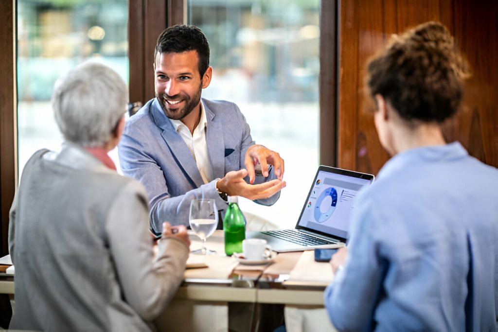 Three people sit at a table in a meeting; one man gestures while speaking, a laptop displays a pie chart from a 13-Week Cash Flow Forecast, and glasses of water and documents are on the table.