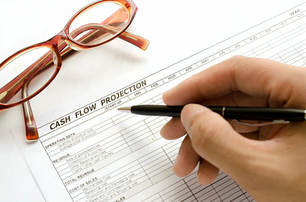 A hand holding a pen reviews a 13-Week Cash Flow Forecast sheet on a desk, with a pair of eyeglasses resting nearby.