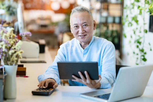 An older man sits at a desk with a laptop, holding a tablet in one hand and using a calculator with the other, smiling at the camera as he reviews his tax deductible expenses.