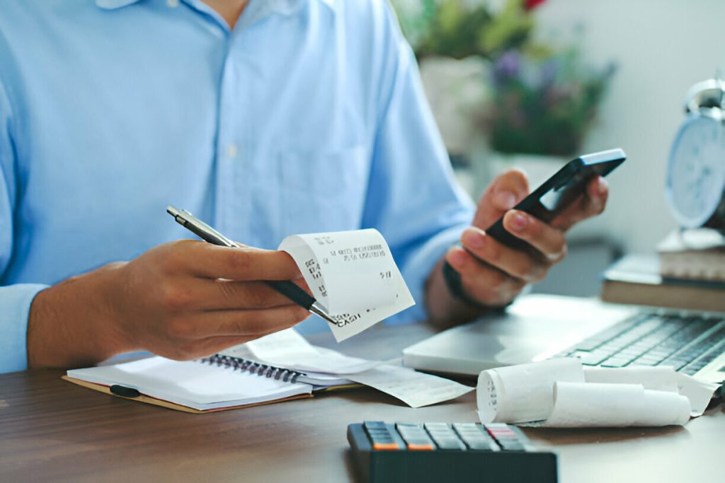 A person sits at a desk holding a receipt and a smartphone, surrounded by a notebook, receipts, calculator, and laptop as they review tax deductible expenses.