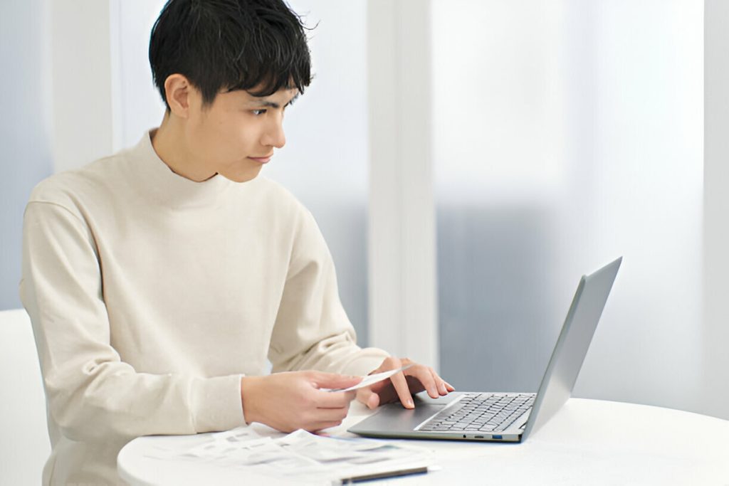 A person sits at a table using a laptop, holding a card and looking at the screen, with several papers related to tax deductible expenses spread out in front of them.