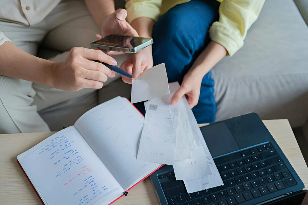 Two people review receipts, use a smartphone, and write in a notebook beside an open laptop on a table as they discuss different types of budgets.