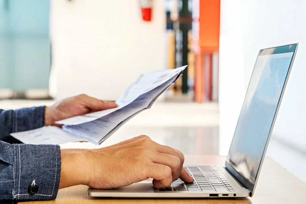 Person holding documents related to types of budgets in one hand while typing on a laptop with the other, seated at a desk in a well-lit indoor setting.