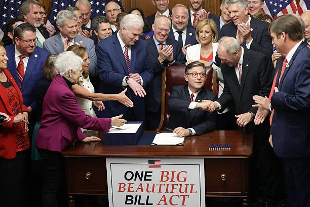 A group of officials and lawmakers shake hands and applaud around a desk with a sign reading "One Big Beautiful Bill Act," celebrating the passage of the One Big Beautiful Bill during a vibrant bill signing event.